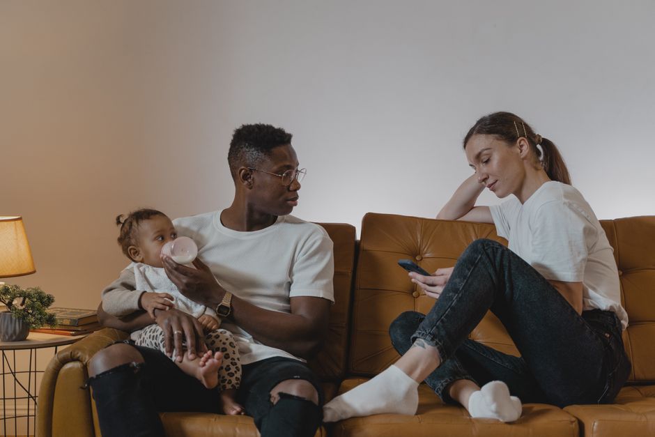 Parents relaxing on a sofa while feeding their baby, enjoying family time with a cellphone.